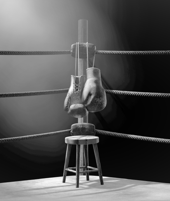 Black and white image of a pair of boxing gloves hung in the corner of a boxing ring, with a stool placed underneath